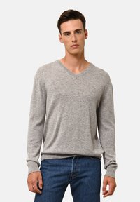 Jasnoszary sweter w serek wykonany z miękkiego materiału, z prążkowanymi mankietami i dołem. Noszony na niebieskich dżinsach, prezentujący luźny fason.