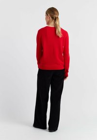 Pull en laine rouge à manches longues et poignets côtelés, associé à un pantalon large noir, vu de dos. Détail de logo subtil sur le pull.