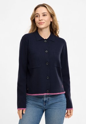 Frau mit welligem blondem Haar trägt eine marineblaue, geknöpfte Strickjacke mit Fronttaschen und pinkem Besatz, kombiniert mit hellblauen Jeans.
