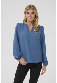 Blaue Bluse mit langen Ärmeln aus strukturierter Stoff; verfügt über einen V-Ausschnitt und geraffte Manschetten, kombiniert mit schwarzen Hosen.