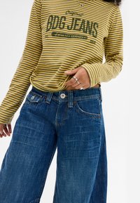 Raidallinen pitkähihainen toppi keltaisen ja vihreän sävyissä, jossa on "BDG JEANS" -teksti. Sen kanssa on yhdistetty siniset farkut, joissa on rento malli ja klassiset taskut.