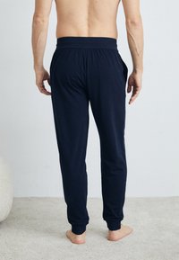 Tummansininen joustava vyötärö sweatpants, joissa on sivutaskut, kapeat lahkeet ja pehmeä kangas, kuvattuna takaa. Paljasjalat vaalealla matolla.