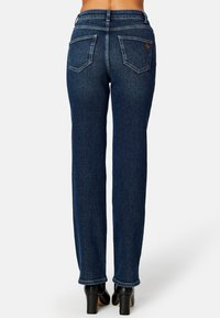 Tmavě modré džíny s rovnými nohavicemi a středním pasem, s dvěma zadními kapsami a kontrastním šitím, nošené s černými vysokými podpatky.