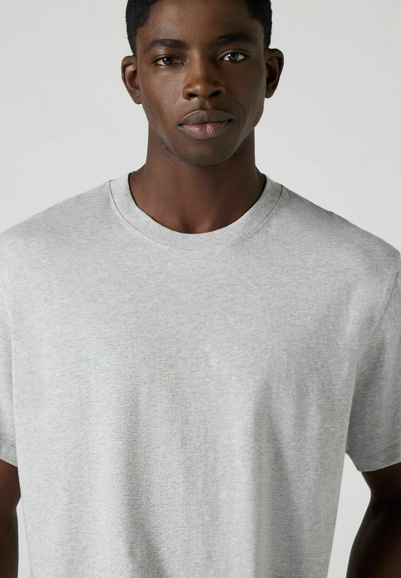 Graues Baumwoll-T-Shirt mit Rundhalsausschnitt, kurzen Ärmeln und weicher Textur, präsentiert auf einem Modell vor neutralem Hintergrund.