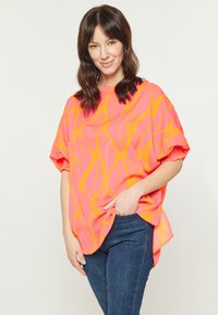 Maglietta oversize arancione brillante con motivo a foglie rosa, maniche corte e orlo arrotondato. Abbinata a jeans blu scuro.