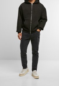 Sudadera negra con cremallera y corte relajado, combinada con unos vaqueros gris oscuro. Zapatillas blancas y beige completan el conjunto. Diseño sencillo, sin patrones visibles.