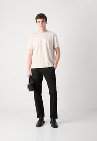 Light beige t-shirt with a subtle logo, paired with black pants and black shoes. The model holds a black bag, standing against a neutral background.