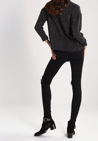 Femme debout, les mains sur les hanches, portant un pull gris foncé, un jean skinny noir déchiré et des bottines noires avec des boucles argentées.