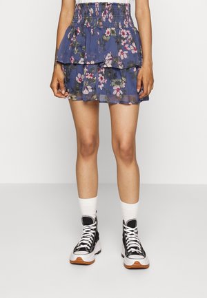Kvinde iført en blå blomsterprintet lagdelt miniskørt, hvide sokker og sorte-hvide høje sneakers, der står på en ensfarvet baggrund.