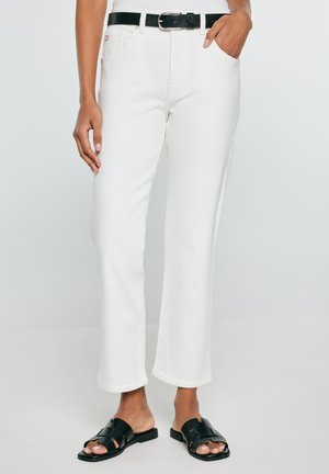 Inimene, kes kannab sirge lõikega valgeid pükse, musta vööd ja musti avatud varbaga libisevaid sandaale, seisab ühe käega taskus.