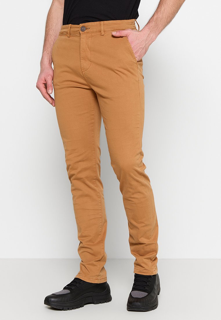 Beżowe płócienne chinosy o slim fit, z zapięciem na guzik i zamek błyskawiczny, kieszeniami bocznymi oraz gładką teksturą. Noszone z czarnymi butami.
