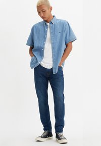 Mlad moški s kratkimi blond lasmi, oblečen v svetlo modro denim majico s kratkimi rokavi, belo T-majico, modre kavbojke in črne superge s poudarjenim gležnjem.