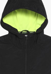 Černá lehká bunda s kapucí, vybavená neonově zelenou vnitřní podšívkou a středním zipem s černými táhly.