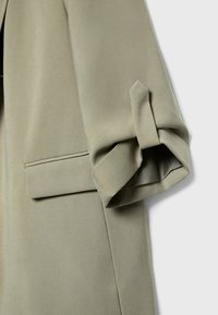 Manche de manteau vert olive clair cintré avec un revers plié et une poche à rabat horizontale sur le côté.