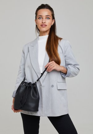 Borsa a secchiello in pelle nera con chiusura a coulisse e accessori in argento, tenuta in mano, abbinata a un blazer grigio e a un top bianco.