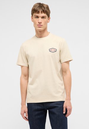 Junger Mann trägt beige kurzärmliges T-Shirt mit kleinem "Mustang Jeans Co" Logo auf der Brust und dunkelblaue Jeans vor einfachem Hintergrund.