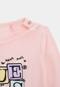 Helles rosa Baumwollhemd mit kurzen Ärmeln und Druckknopfverschluss, das auf der Vorderseite bunte Blockbuchstaben und ein verspieltes Textdesign zeigt.