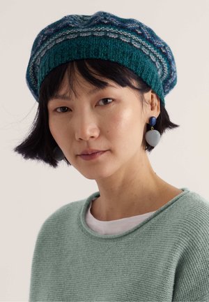 Femme aux cheveux courts noirs portant un béret en tricot vert à motifs, des boucles d’oreilles pendantes bleu-gris et un pull vert clair sur une chemise blanche.