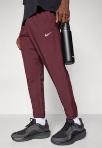 Burgundijas krāsas Nike sporta bikses ar rāvējslēdzēju potītēs, apvienotas ar melnām Nike kedām un melnu ūdens pudeli, kas turēta rokā.