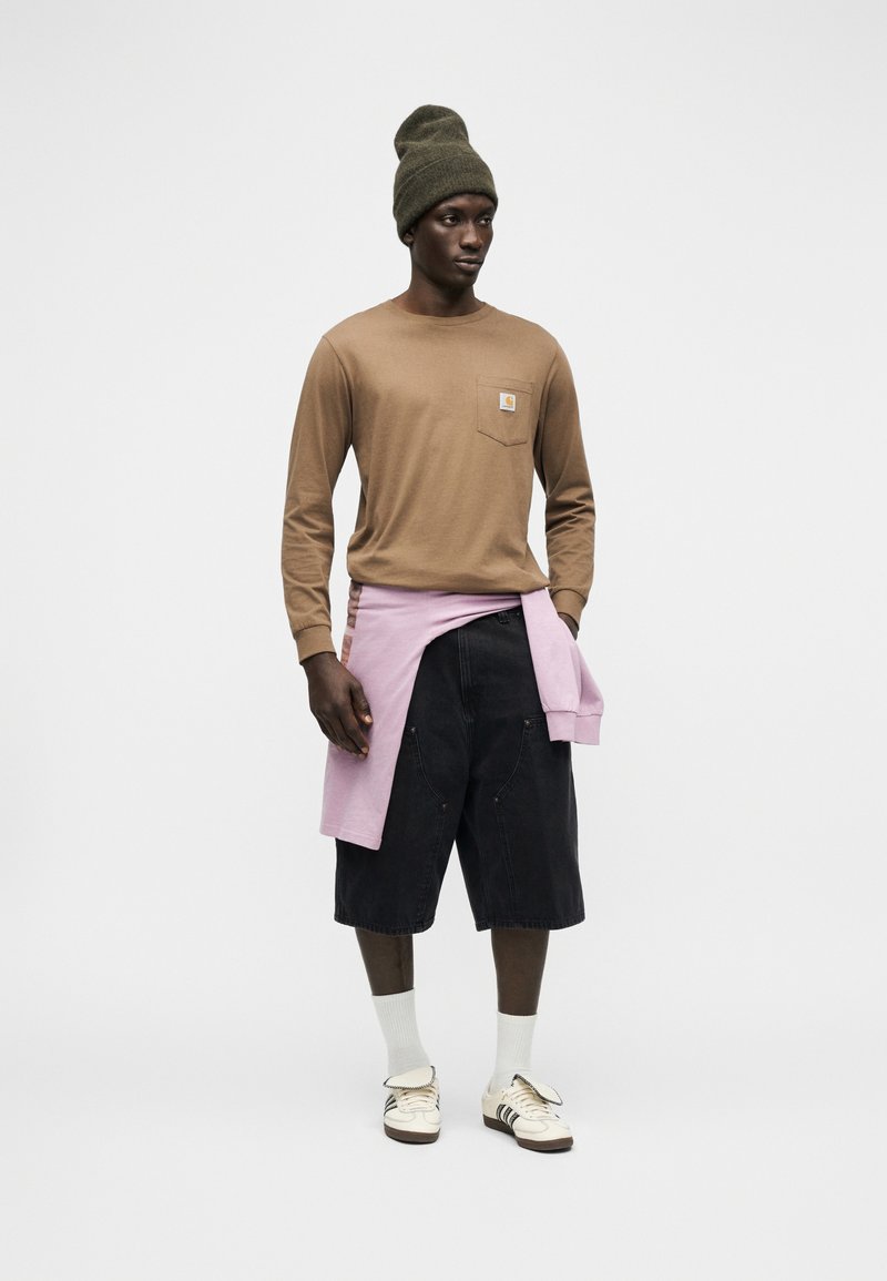 Uomo che indossa un berretto oliva, una camicia marrone a maniche lunghe, pantaloncini neri fino al ginocchio, calzini bianchi e scarpe da ginnastica con una felpa rosa legata in vita.