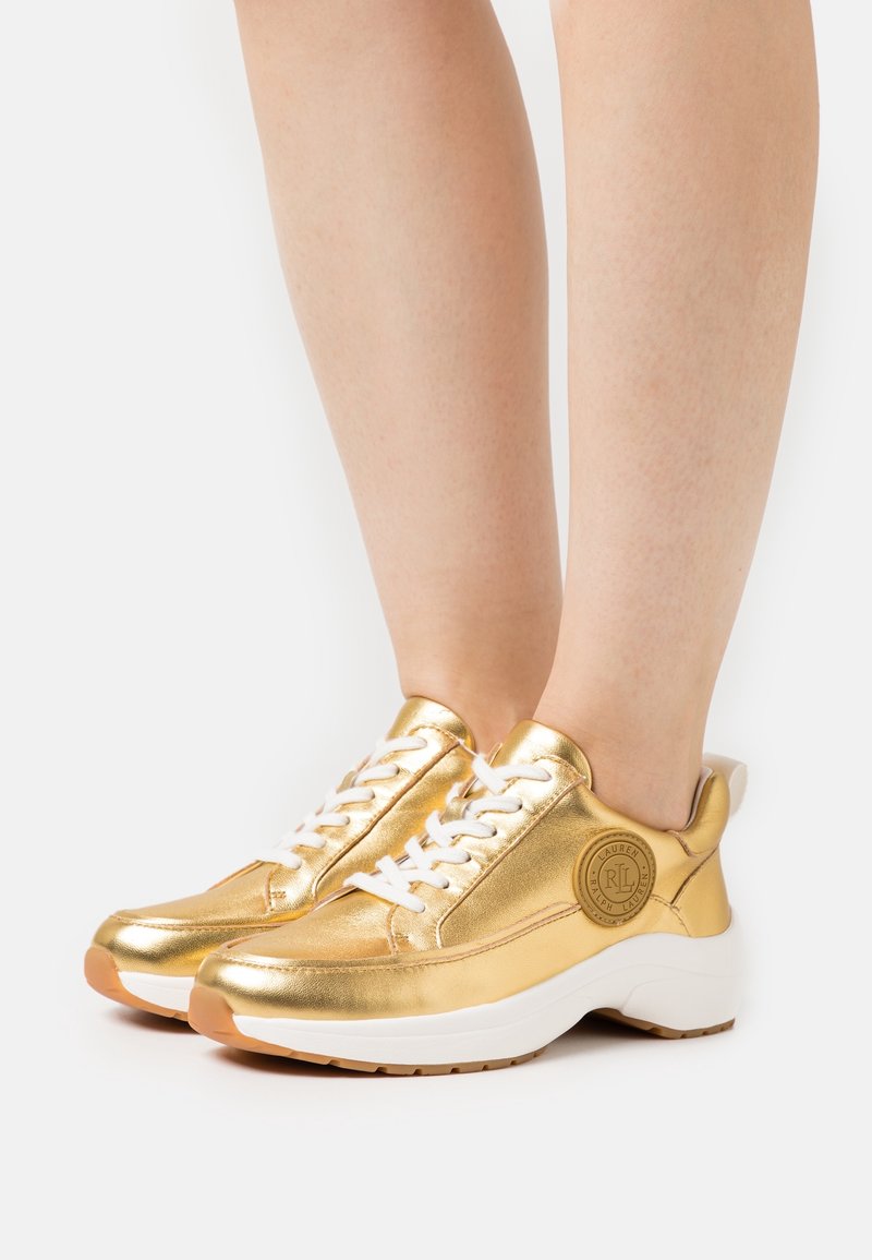 Guldmetalliska sneakers med vita snören, rund tå, logodetalj på sidan och en tjock vit sula med brun yttersula.