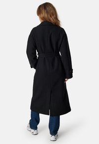 Cappotto lungo nero realizzato in un misto lana, con vita regolabile, aperture laterali e ampi revers. Abbinato a jeans blu e sneakers.