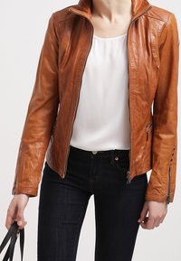 Giacca in pelle marrone con design aderente, colletto alto e chiusura frontale con zip. Presenta polsini con zip e dettagli cuciti discreti.