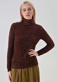 Maglione dolcevita marrone realizzato in materiale morbido e soffice. Presenta maniche lunghe e una vestibilità comoda, con un leggero effetto lucido su tutto il tessuto.