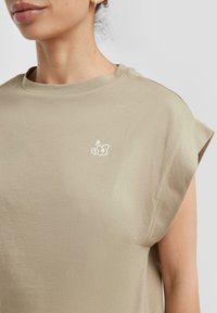 Chemise beige clair à manches courtes avec un col rond et un petit motif brodé. Texture lisse avec une coupe décontractée et un léger éclat.