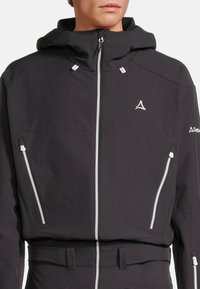 Giacca softshell nera con chiusura a zip frontale, due tasche laterali e cappuccio. Presenta dettagli in zip bianchi e logo ricamato sul petto.