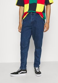 Jeans de mezclilla azul con un corte relajado, con pasadores para cinturón y un cierre de botón único, combinados con zapatillas negras y una blusa multicolor.
