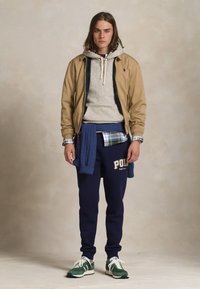 Beige bomber dzseki egy szürke kapucnis pulcsi fölött, egy kockás inggel rétegzett. Tengerészkék melegítőnadrág felirattal, zöld tornacipő. Kényelmes, réteges öltözet.