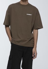 Camiseta de algodón marrón de corte oversized con mangas cortas, que presenta un pequeño logo blanco en el pecho y un cuello redondo.
