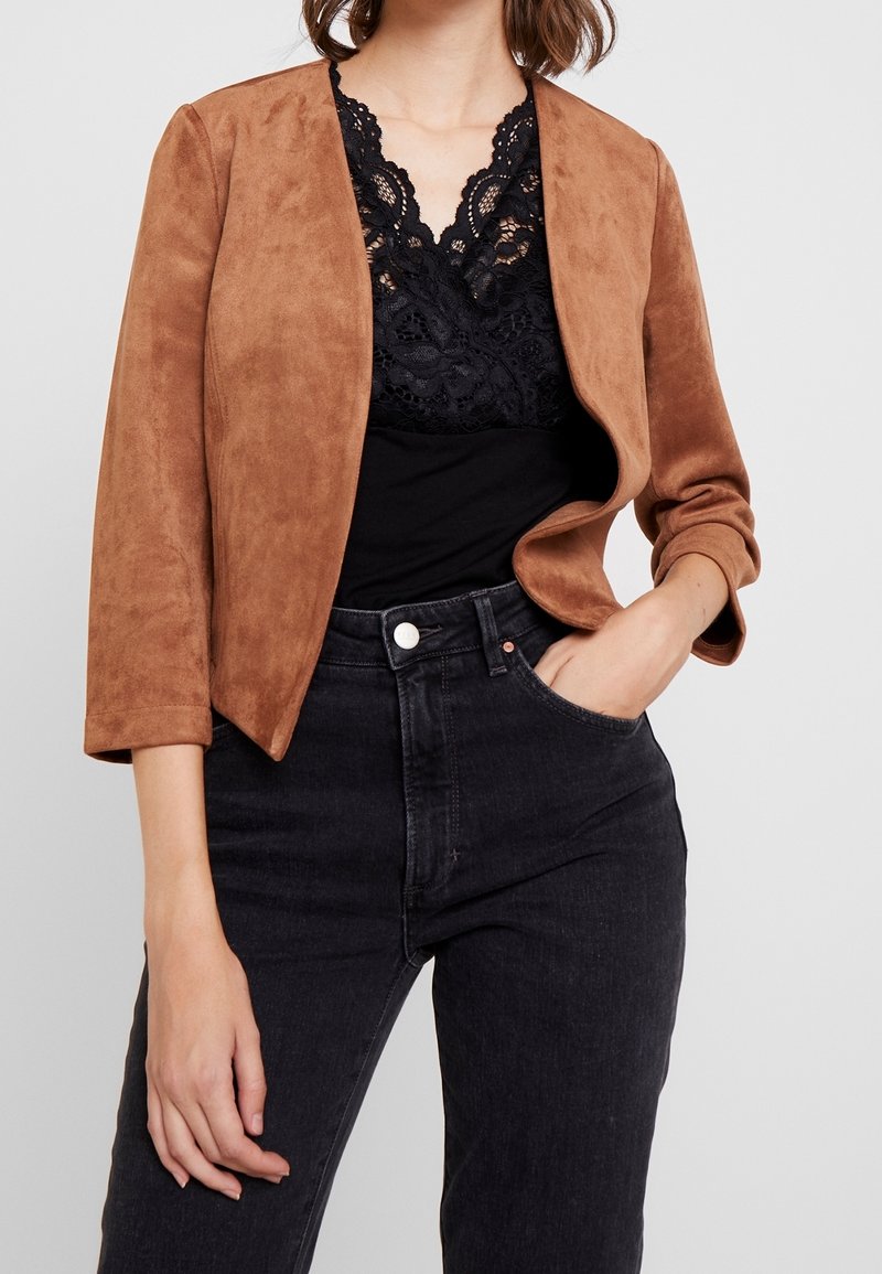 Femme portant une veste courte marron sur un top en dentelle noire et un jean noir taille haute, une main dans la poche, sur un fond uni.