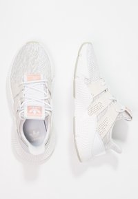 Valkoiset urheilukengät, joissa on erilaisia tekstuureja, mukaan lukien verkkoa ja sileitä materiaaleja. Vaaleanpunaiset yksityiskohdat ja säädettävät nauhat. Kevyt muotoilu.