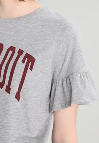 Grå kortärmad skjorta med volangärmar och granatröda bokstäver "OIT" synliga, bärs av en person mot en ljus enfärgad bakgrund.