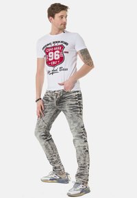 Weißes T-Shirt mit rotem Grafikdesign, graue ausgebleichte Jeans mit detaillierter Stickerei und graue Sneakers mit mehrfarbigen Akzenten.