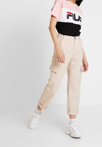 Pantalones cargo beige con múltiples bolsillos, dobladillos con puños y una textura suave. Combinados con una camiseta de logo de manga corta en negro, blanco y rosa.