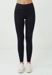 Modella che indossa leggins neri e scarpe da ginnastica bianche si trova contro uno sfondo chiaro e semplice, mostrando un outfit atletico casual.