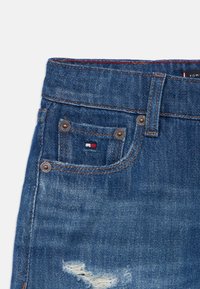 Modri kavbojki iz denim materiala se ponašajo z obrabljeno teksturo in raztrganinami, bakrenimi dodatki ter majhnim tricolor logotipom na sprednjem žepu.