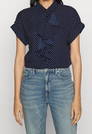 Blouse bleu marine à manches courtes avec pois blancs et détail volanté sur le devant, associée à un jean taille haute bleu clair.