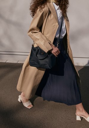 Femme marchant portant un trench-coat beige, une chemise blanche, une jupe plissée bleu marine, des sandales blanches à talons blocs et portant un sac à main noir.