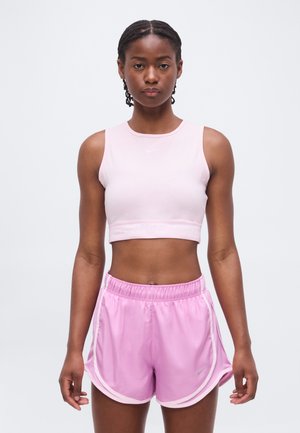 Kobieta stojąca przodem, ubrana w jasnoróżowy bezrękawnik typu crop top i pasujące sportowe szorty na jednolitym białym tle.