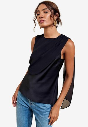 Femme portant un haut en satin noir sans manches avec un détail drapé et un tissu transparent drapé dans le dos, associé à un jean bleu et des boucles d'oreilles dorées.