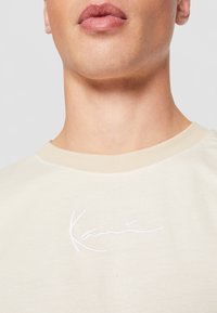 Beige sweatshirt with a crew neckline, featuring a white embroidered logo. The fabric appears smooth with a soft texture.