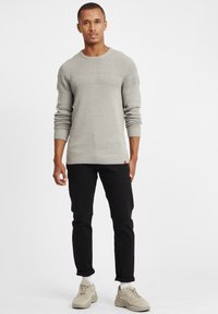 Hellgrauer Strickpullover mit strukturierten Ärmeln und ribbeltem Saum, kombiniert mit schmalen schwarzen Jeans und beigen Sneakern.