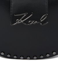 Černá kožená kabelka s metalickým logem 'Karl', se stříbrnými nýty podél dolního okraje a hladkým povrchem.
