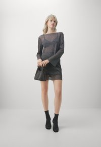 Vestito mini lungo nero trasparente con texture scintillante, scollo a barca e maniche a campana. Abbinato a una borsa a tracolla nera coordinata e calze.