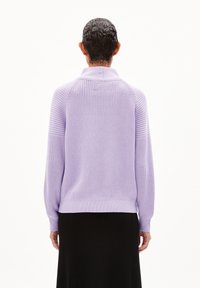 Lila neuletakki ribbitekstuuri, korkea kaulus ja laskeutuvat hartiat, yhdistetty mustan hameen kanssa, näkyy takaa vaaleaa taustaa vasten.