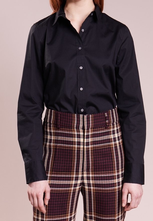 Femme portant une chemise noire à manches longues boutonnée, rentrée dans un pantalon taille haute à carreaux bordeaux et blancs, sur un fond uni.